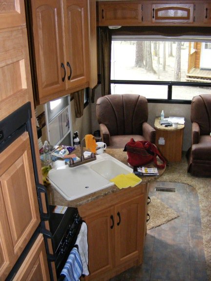 The two-basin sink and its cabinet sticks out          into the living area at about a 45 degree angle. A small amount of          the range and the refrigerator can be seen at the left side.           The two easy chairs are visible near the window at the back of the          room.