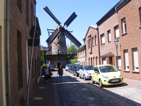 At the end of a narrow street, a 17th century windmill stands.            The street is lined with brick buildings and cars are parked along            one side. 