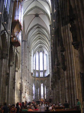 Looking toward the end of the cathedral from the center,            the interior appears narrow, flanked by vertical columns           rising to the pointed arches of the ceiling high above.           The sides appear dark while the ceiling and end are light.