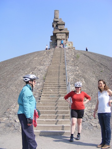 Gail in a blue shirt and bike helmet, Ingrid in a red shirt and             bike helmet and Katrin in a white shirt are standing at the bottom             of a long stairway that ascends a hill of rocks. At the top of the             hill is a sculpture that consists of massive blocks stacked into             a tower that is about 30 feet high. 
