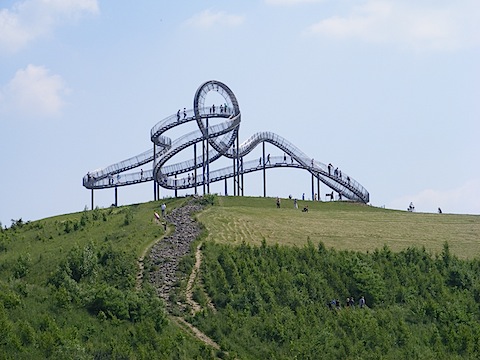 At the top of a grassy mound is a curlicue structure, maybe 130 feet          across and 40 feet high. People are walking on the pathways formed by          the structure.