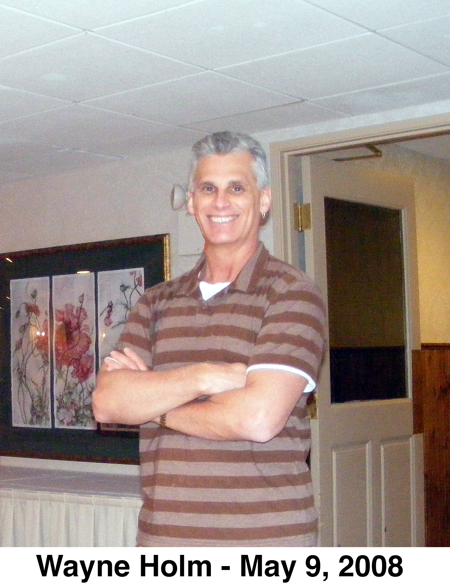 The photo shows Wayne standing with his arms folded in front of
           a door and a wall with paintings.