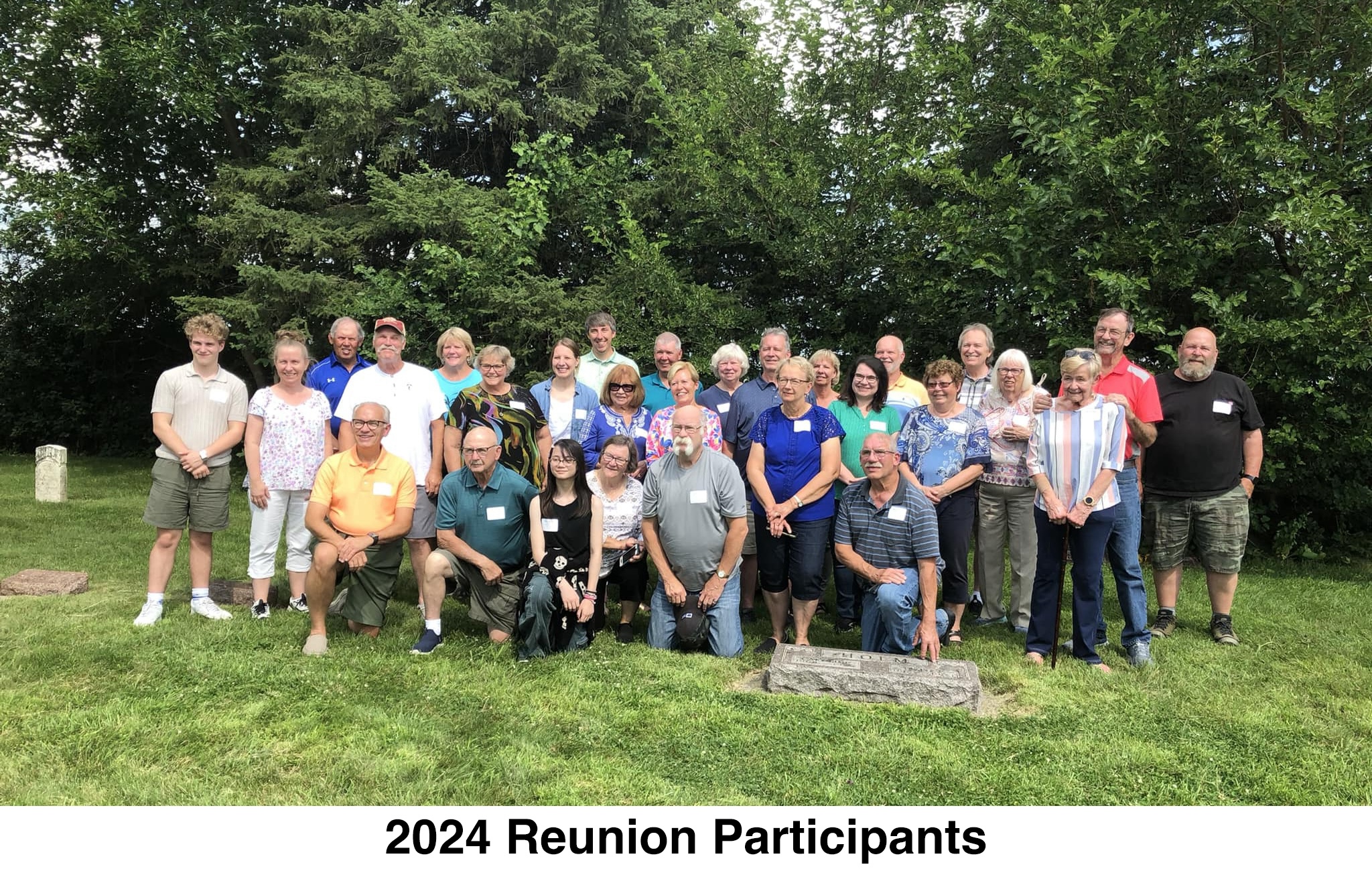 There are three rows of people. The front row is kneeling. 
       The Holm grave stone is in front of them and a row of trees is behind them.