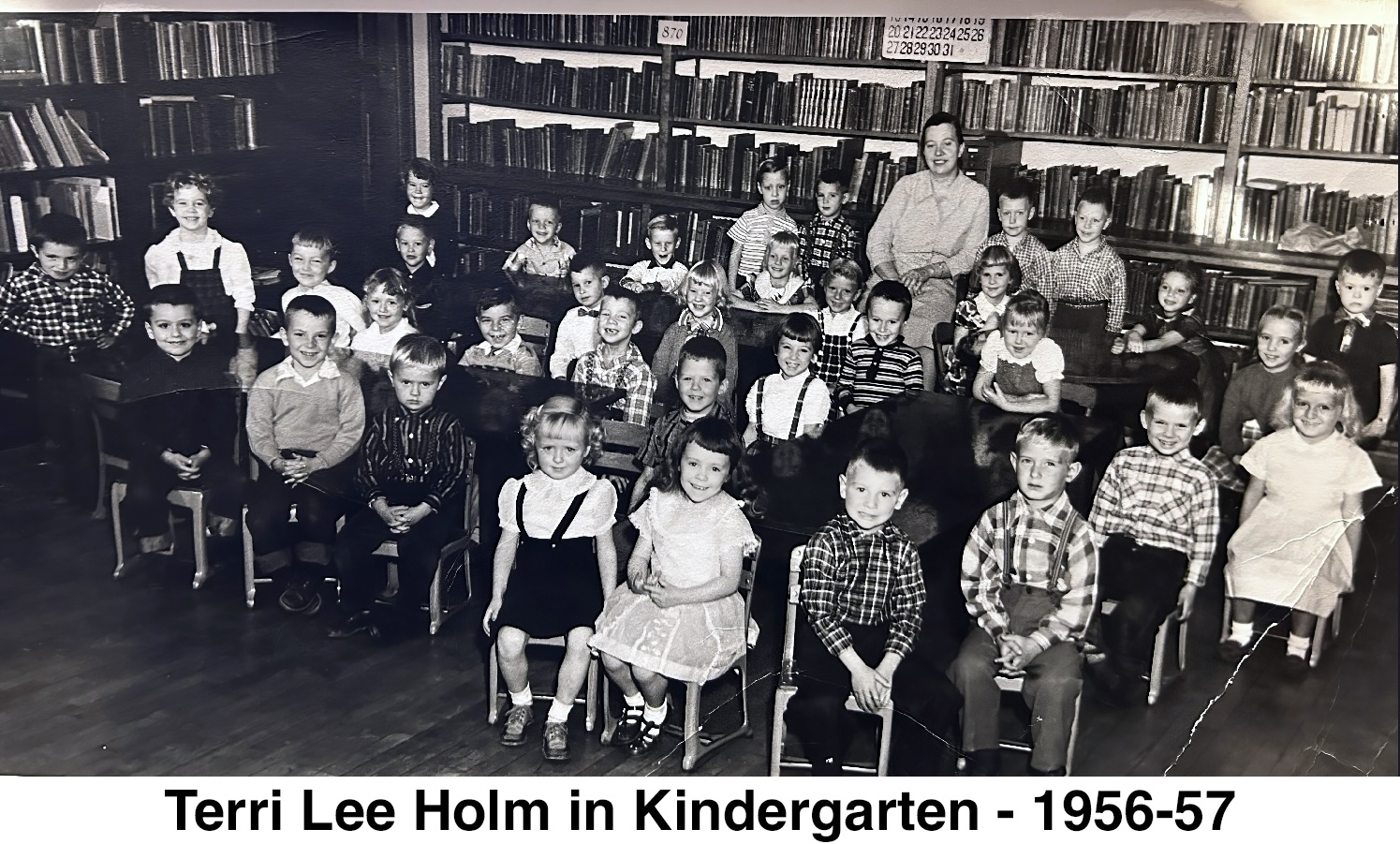 A black&white photo of 35 students and their teacher in front of a wall of books.
       Most are sitting and smiling at the photographer. 