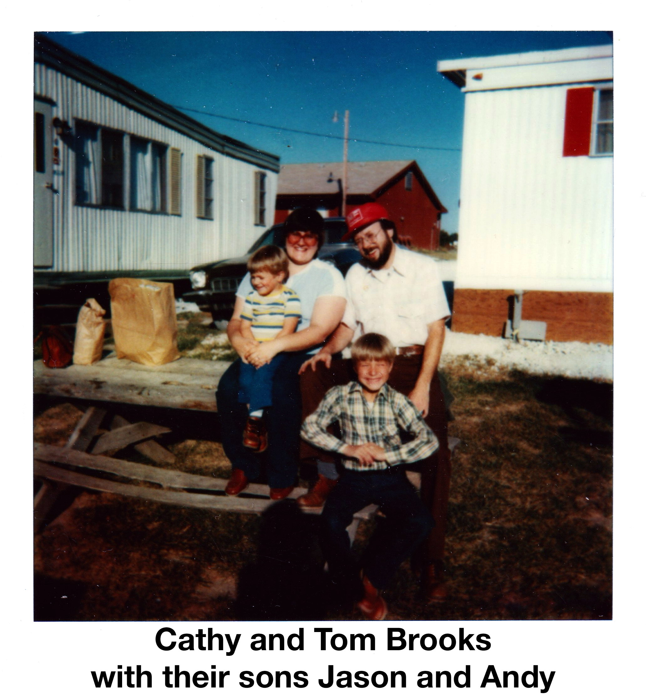 The family is sitting on a picnic table between mobile homes on
            a sunny day