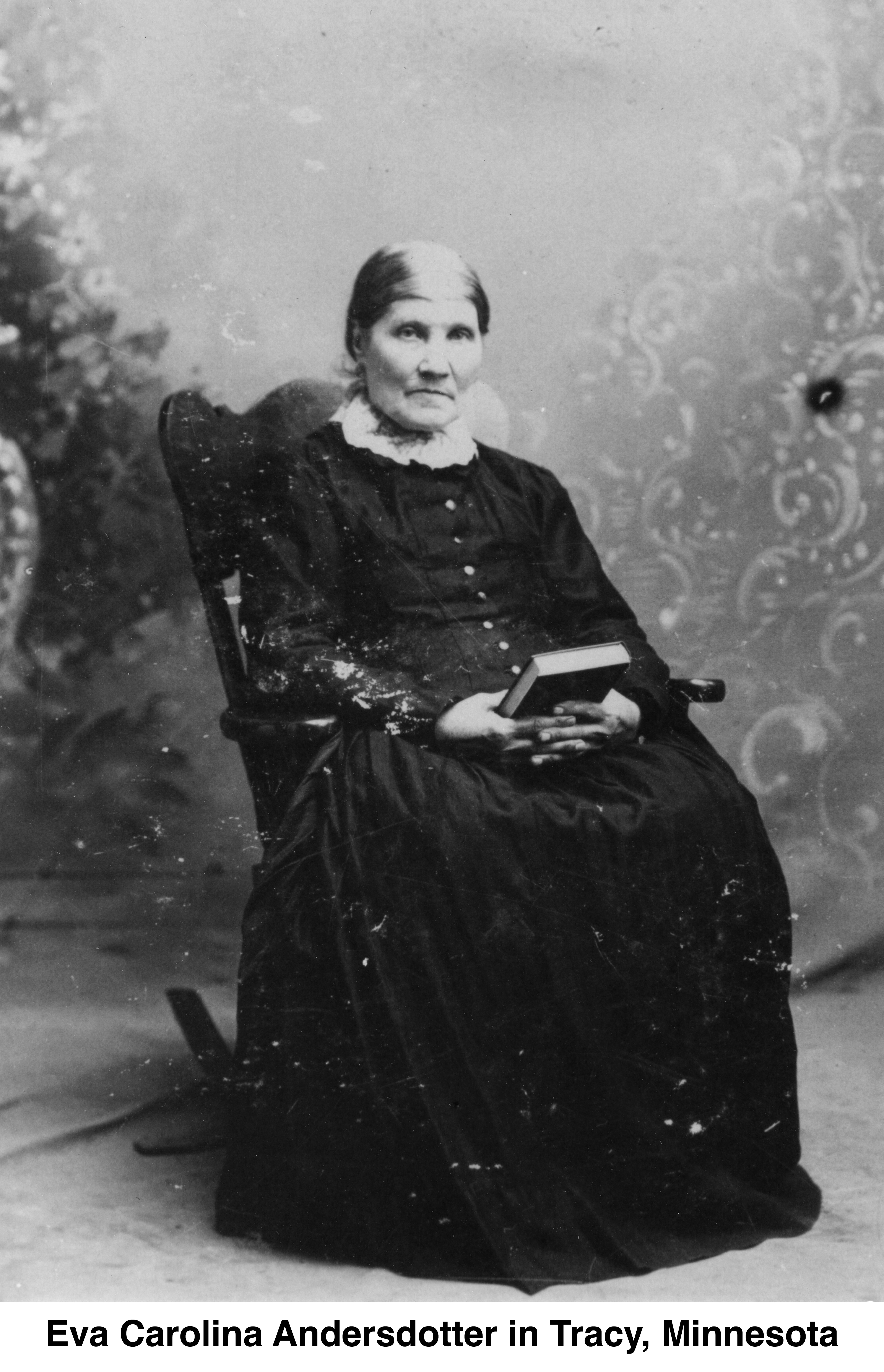 She is sitting in a rocking chair, holding a Bible and wearing a 
         dark dress.