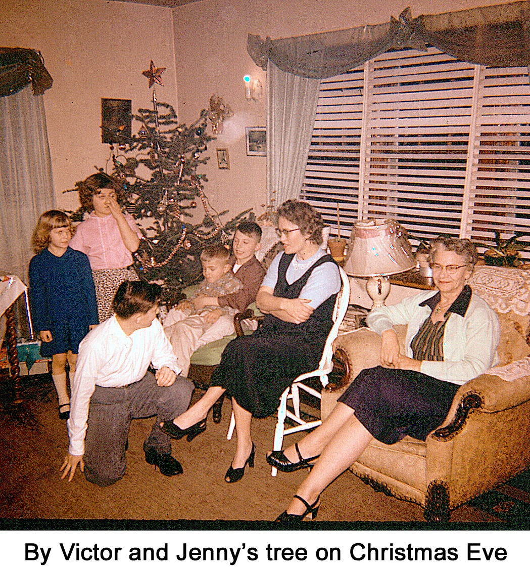 The tree is in the corner with children and adults standing and sitting with 
    their backs to it.