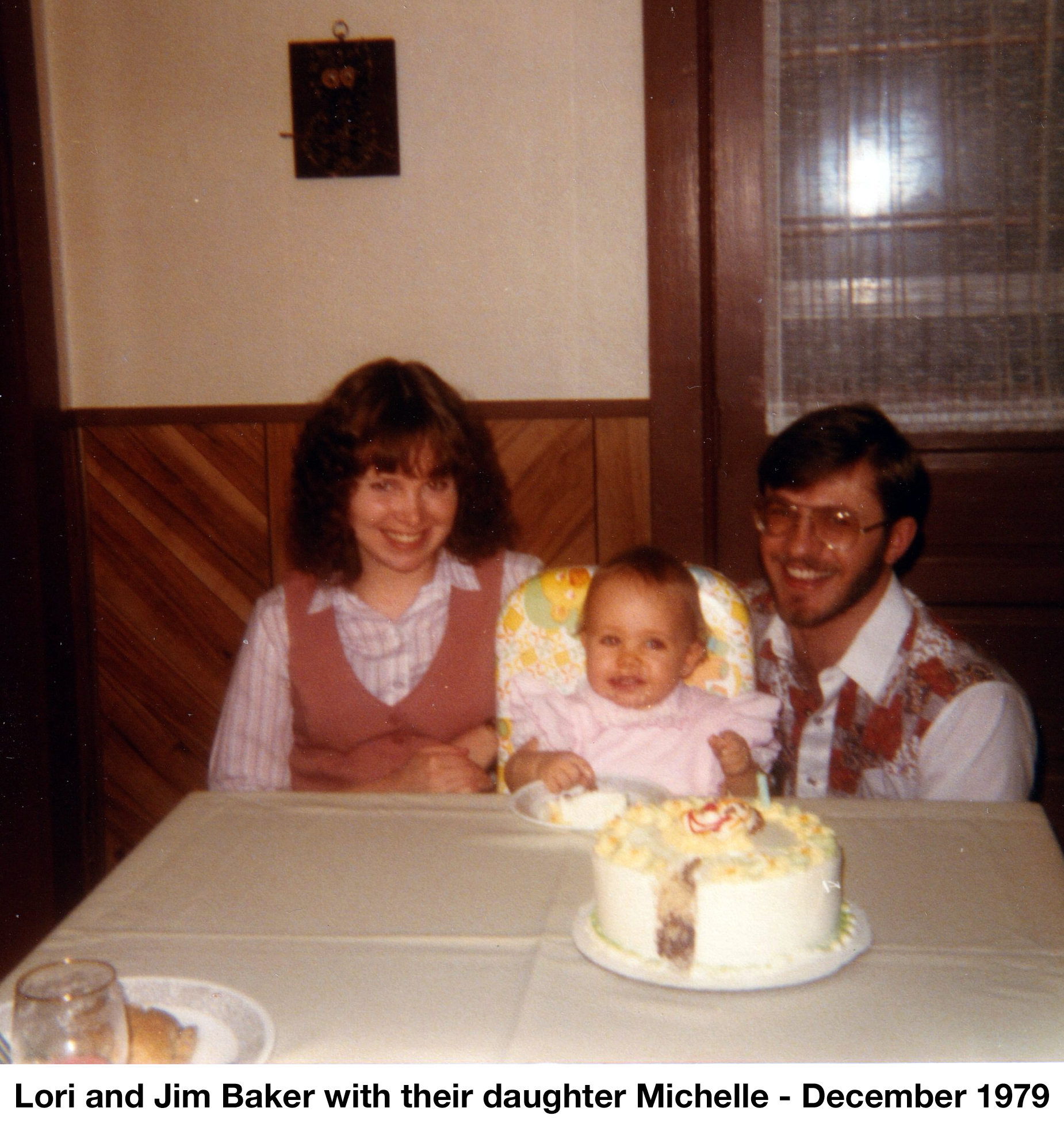 <Michelle is in a highchair with her parents on either side and a 
         cake on the table in front of her