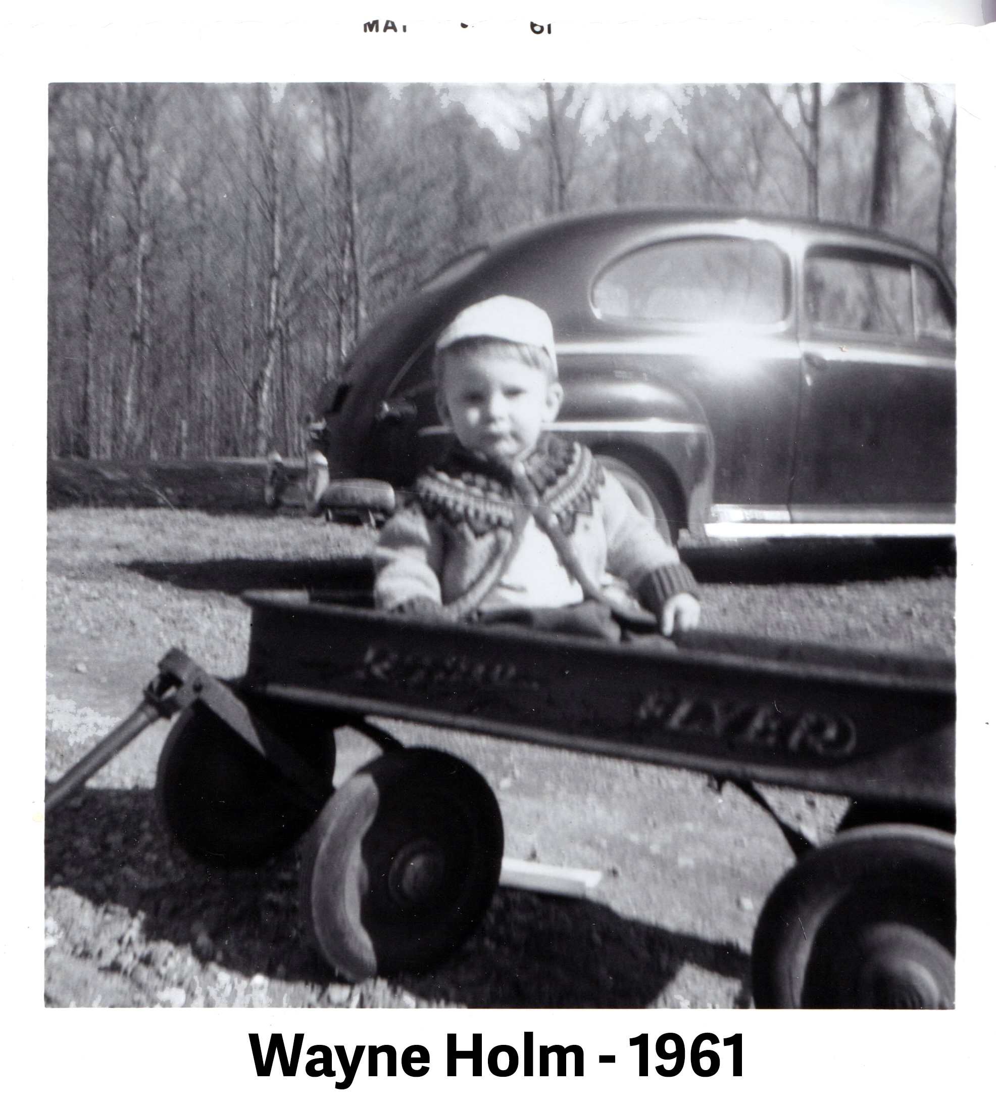 Wayne is wearing a sweater and sitting in an American Flyer wagon.
              A 1940s-era, 2-door car is behind him.