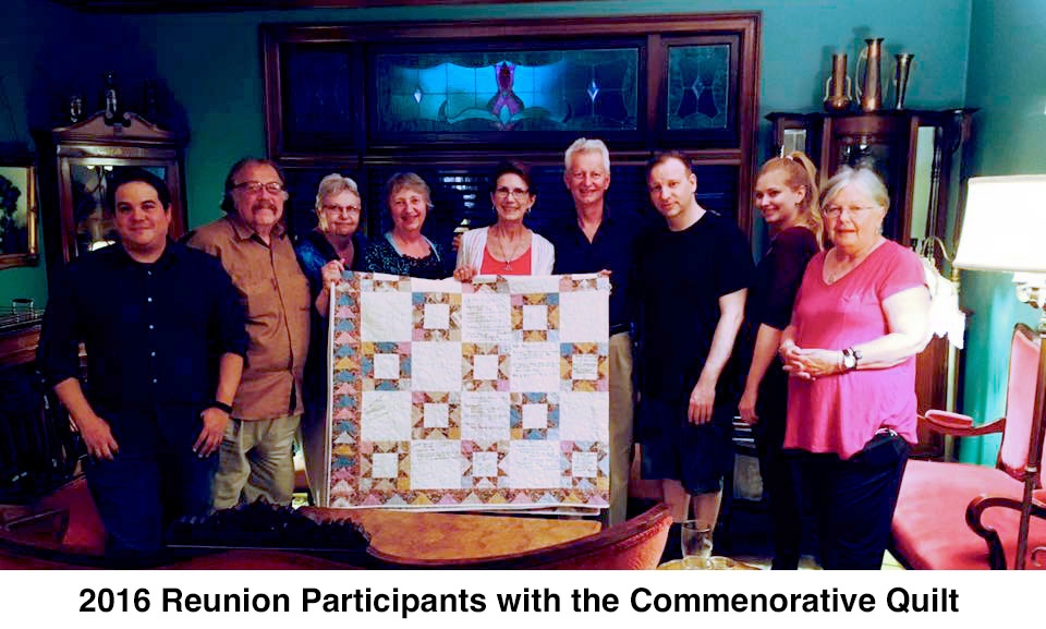 Nine participants are standing in a room with the quilt held in front of them