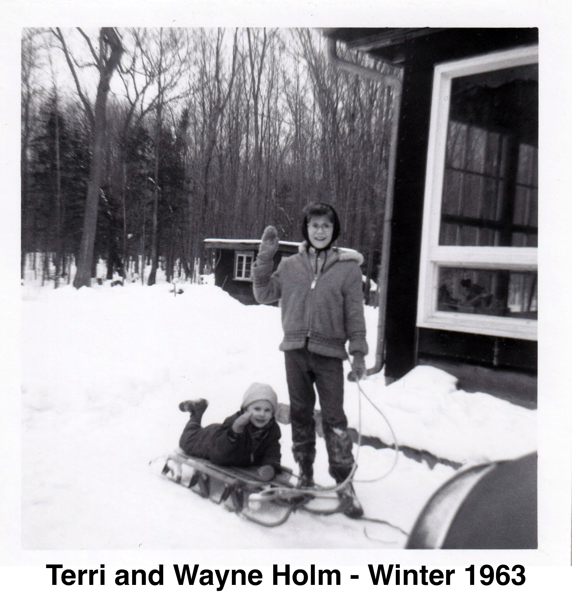 Wanye is lying on a sled and Terri is hold its rope. Both stand 
            along side the house with the trees and shed visible in the background
