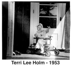 Terri sits in a walker with the door behind her. 
           She is reaching toward the photographer. 