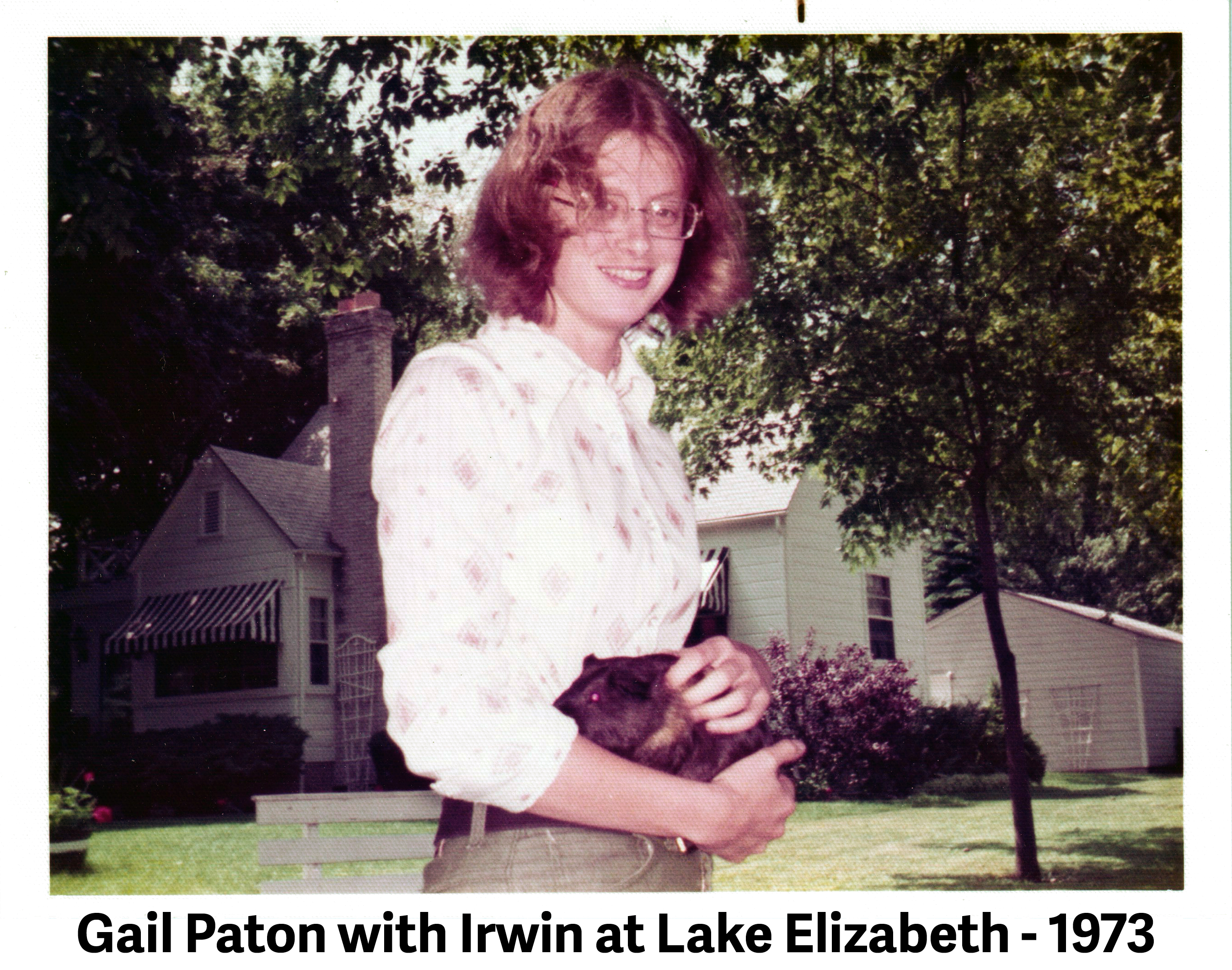  Gail is standing behind the house and holding her guinea pig 
                by her waist.  