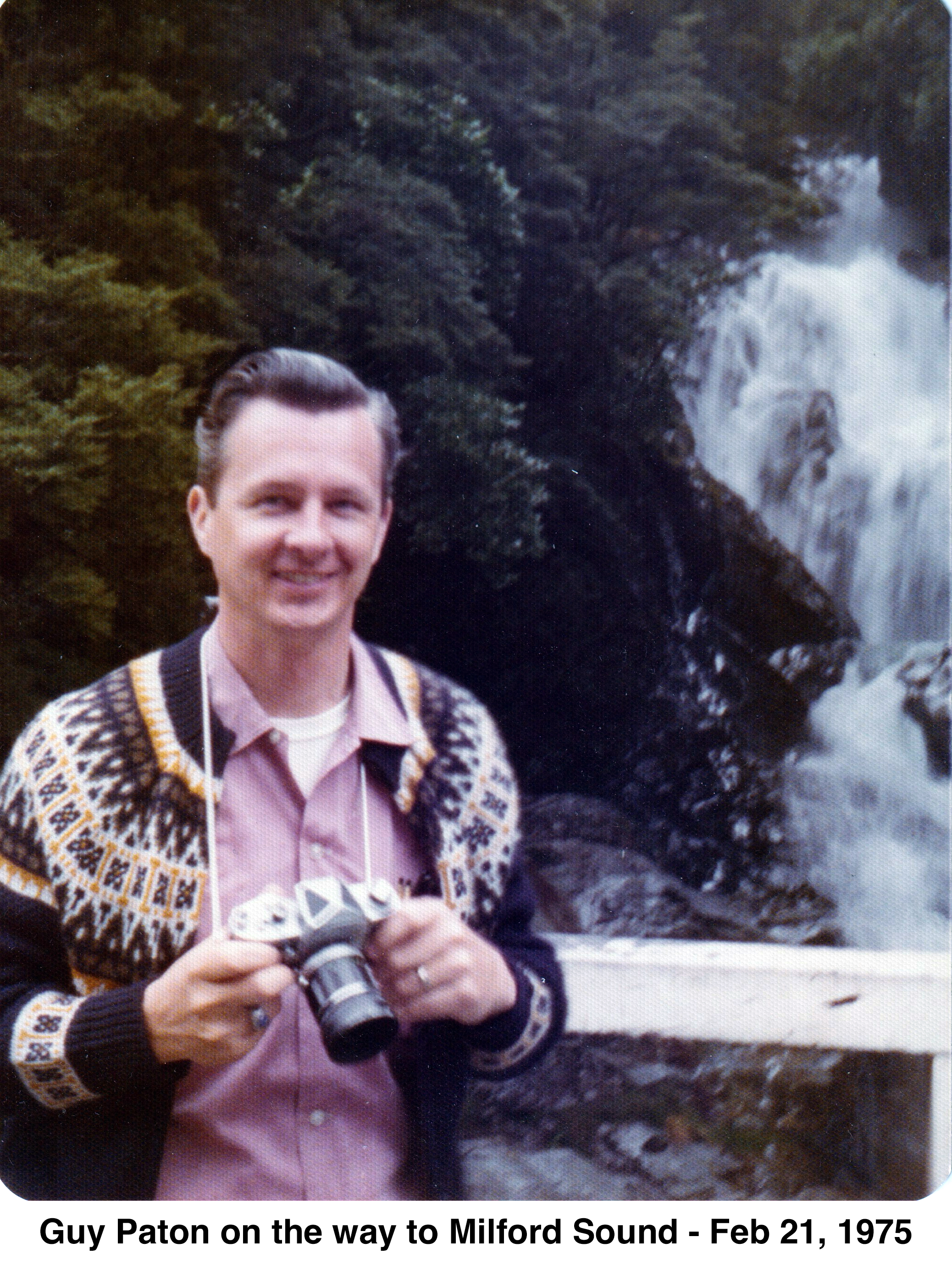 Guy is wearing a sweater and holding a camera in front of green 
              bushes and a waterfall