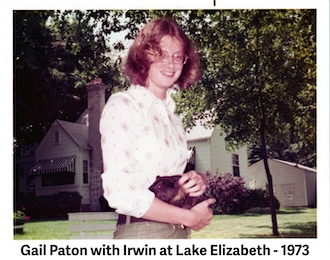 Gail is standing behind the house and holding her guinea pig 
                by her waist. 