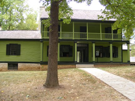 The house is              painted green. The main part is two stories              with a covered porch and a covered balcony               above it. To the left is a one-story wing              with no porch.