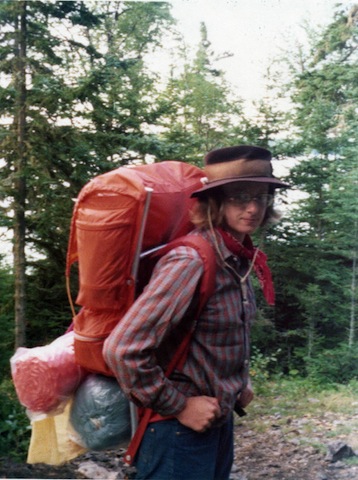 Gail wears a long-sleeved shirt and a brown rimmed hat, and carries an orange backpack with a blue sleeping bag, an orange bubble mattress and a yellow rain coat hanging below the pack.