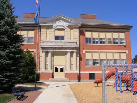The twow-story brick front of the old school. The entrance
               is elaborate with pillars. 
               An evergreen tree is on the left and a playground on the right.
               The sky is very blue.