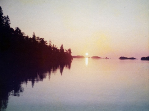The sun is seen risen over the water with a tree-covered              shoreline silhouetted to the left and some small islands               silhouetted to the right.