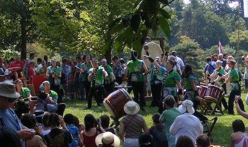 Many drummers in green shirts      are surrounded by an audience