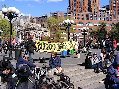 Two men hold a        yellow banner proclaiming Occupy Wall Street. Two men in        the foreground play chess. The sky is a beautiful blue.