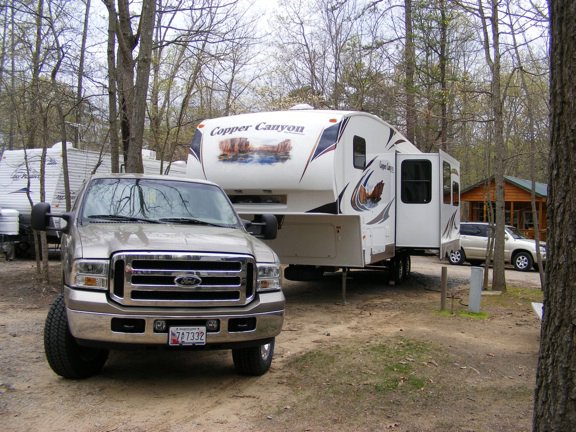 Our tan Ford F-250 is parked           in front of the Copper Canyon trailer. Leafless trees           surround the area.