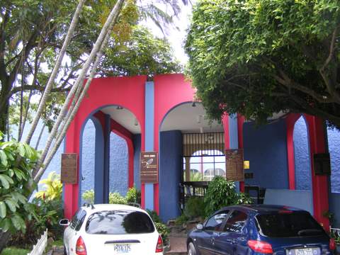 An entrance into the red and blue painted building,                     with cars parked in front and trees
