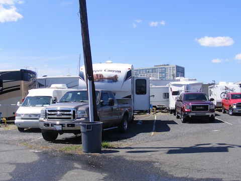 Our Ford F-250 truck and Copper Canyon 5th wheel trailer 
               are parked behind a light pole in the Liberty Harbor RV Park. 
               The ground is covered with asphalt and gravel. Campers are 
               very close together side-by-side and back to back. The
               sky is blue with a few puffy white clouds. 