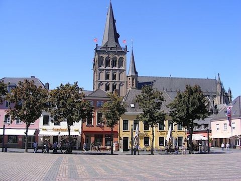 Across a wide, paved open area stand a line of colorful shops.                 Rising high above the shops is the bell tower of the cathedral.