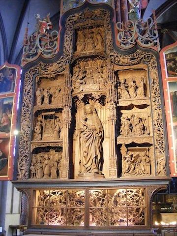 A large panel of Mary with baby Jesus is in the center of this            carved, wooden panel. This is surrounded by seven smaller depictions            of religious scenes. Ornate scrollwork surrounds them all.
