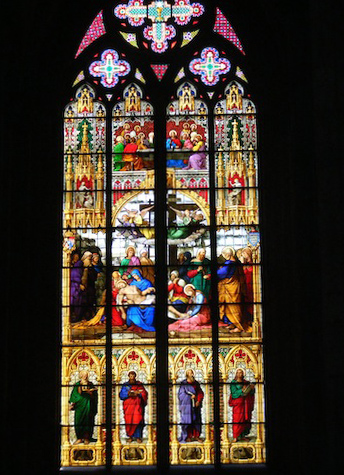 This large colored window incorporates many images. At the           bottom in an area four panels wide and three high are representations           of the writers of the four gospels standing in archways. Each           has a representative animal at his feet. Above them in an area           four panels wide and four high is a representation of the body           of Jesus being removed from the cross. Above that is a two by two           picture of people around a table, possibly a representation of            the last supper. Above that is an area with two smaller and one           large cross, mostly in the colors of lavender, rose and white.