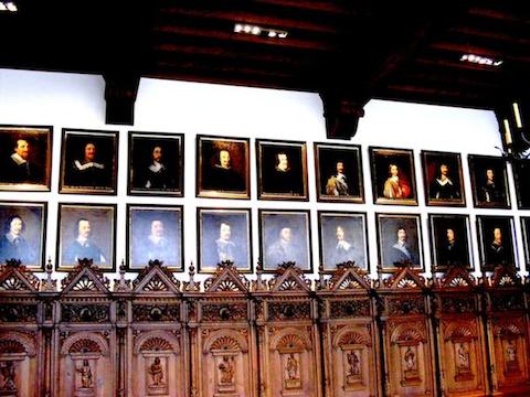 Twenty portraits of men in two rows are seen. Most of the men are             wearing black garments with a collars. The young Louis XIV of France              has a gold and crimson garment. Below the portraits can be seen             the ornately carved wooden backs of seats that line the wall.