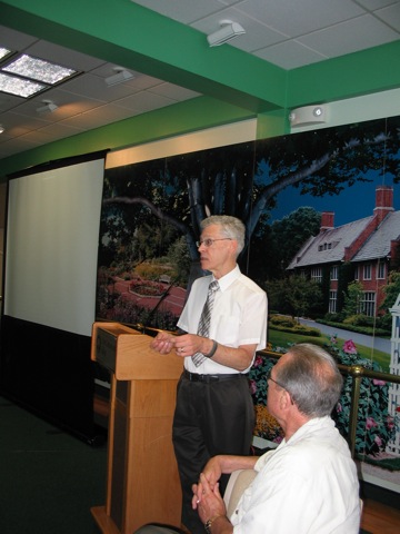 Al standing near a podium. On the left of
               the photo is a projection screen. On the right
               the back of a man's head, listening to the talk.