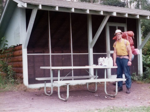 Al stands next to a picnic table and in front of the screen-front shelter, and carries an orange backpack with a blue sleeping bag and an orange bubble mattress hanging below the pack and the orange tent on top of the backpack.