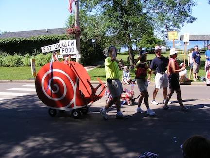 A red-colored cardboard snail on a wagon pulled
               by some walkers has signs for Eat Local and Slow Food