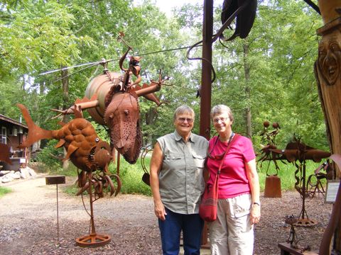 Linda and Gail are standing with the Attack Dragon
               in the background. The Dragon has its head lowered
               and has a helicopter-like rotor spinning on its back.
               Near the right side of the photo is a deep-sea looking
               creature. All the creatures are rusty. Trees are in the
               background.