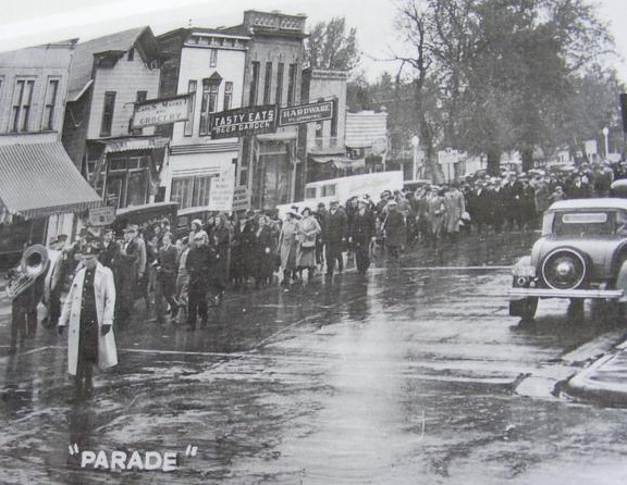 A black and white photo showing a parade in front of
               the west side of Freeman Street on October 25, 1936. People
               are wearing overcoats and the street looks wet as if it
               is raining or has rained recently.