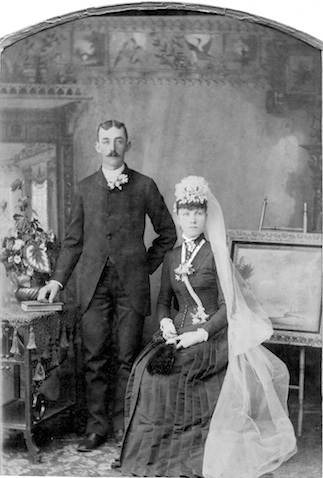 Fred Henjes is standing to the
            left of Ella, who is sitting, possibily on a stool. He is
            wearing a dark suit and she is wearing a long, dark 
            dress. Her hat has a long, white veil attached. 
            There is a table and a painting on an easel in the 
            photograph.